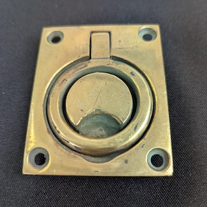 May include: A rectangular brass flush pull handle featuring a central circular ring that pivots within a recessed plate. The metal surface displays a weathered, aged patina with visible scratches and dark oxidation marks around the edges.