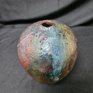 May include: A spherical ceramic vase with a textured, uneven surface. The glaze features a mottled blend of metallic copper, deep blue, forest green, and earthy brown tones. It has a small, circular opening at the top and a rounded body.