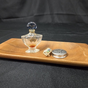 May include: A rectangular wooden tray holding a clear glass Shalimar perfume bottle with a blue stopper, a small gold-tone brooch with blue and green stones, and a round silver-tone metal container with a decorative perforated lid.