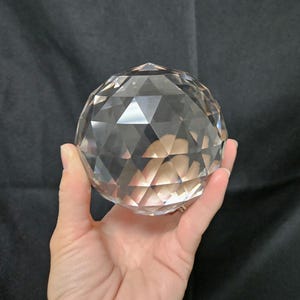 May include: A clear, spherical glass paperweight featuring a multi-faceted geometric surface. The object is transparent and reflects light through its numerous triangular and diamond-shaped cut faces, creating a complex, prismatic appearance.
