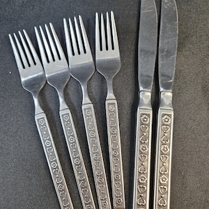 May include: A set of four stainless steel forks and two stainless steel knives. Each piece of flatware features a long, rectangular handle decorated with a repeating embossed pattern of small flowers and heart shapes along the entire length.