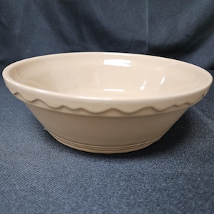 May include: A beige ceramic bowl with a smooth, glossy finish and a wide, circular opening. The exterior features a decorative scalloped edge detail just below the rim and subtle horizontal ridges near the base, creating a simple, functional design.