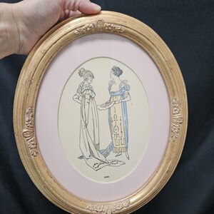 May include: An oval-shaped gold-toned frame with ornate floral detailing, containing a print of two figures in 1810-style attire. The print features a light pink mat and displays the text 1810 at the bottom center, showcasing historical fashion.
