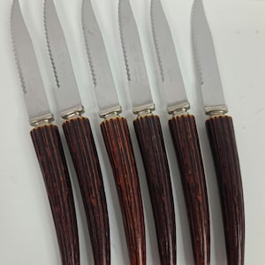 May include: A set of six steak knives with silver-toned serrated metal blades and dark brown, textured, curved handles. Each blade is engraved with the text Sheffield England. The knives are arranged in a parallel row against a plain background.