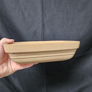 May include: A rectangular baking dish with a smooth, tan-colored ceramic finish. The dish features a wide, flat rim at the top and two distinct horizontal ridges running along the exterior sides, providing a structured and clean appearance.