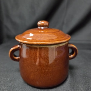 May include: A small, round, brown ceramic pot with a glossy finish, featuring two curved side handles and a matching removable lid with a central knob. The vessel has a smooth, uniform glaze and a simple, functional design for storage or serving.