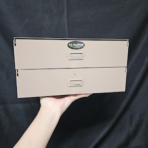 May include: A beige metal two-drawer storage cabinet with a rectangular shape. The top drawer features a black oval label reading KE-MASTER MEDIA, PENNSYLVANIA. Each drawer has a centered metal handle with a slot for a label insert.