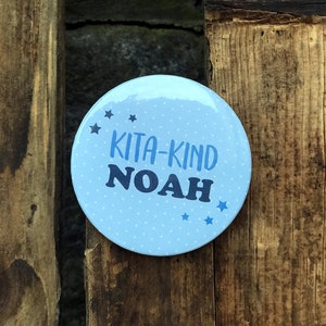 Könnte beinhalten: Ein blauer und weißer Knopf mit weißen Punkten und blauen Sternen. Der Knopf trägt die Aufschrift "Kita-Kind Noah" in schwarzer Schrift.