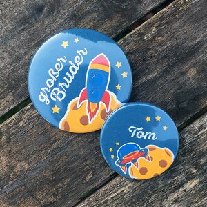 Könnte beinhalten: Zwei runde blaue und gelbe Buttons zum Thema Weltraum mit einer Rakete und einem Raumschiff. Der größere Button sagt "großer Bruder" und der kleinere Button sagt "Tom".