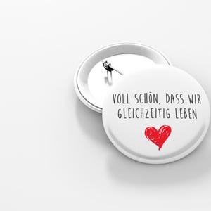 Könnte beinhalten: Weißer Knopf mit einem roten Herzen und dem Text "Voll schön, dass wir gleichzeitig leben" in Schwarz.