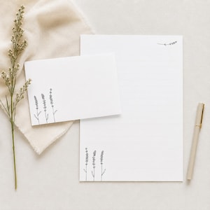 Könnte beinhalten: Ein weißes Schreibwarenset bestehend aus einer rechteckigen Karte und einem linierten Blatt Papier, beide verziert mit schwarzen Strichzeichnungen von Lavendelzweigen. Ein beigefarbener Stift liegt rechts vom Papier und ein getrockneter Lavendelzweig liegt links.