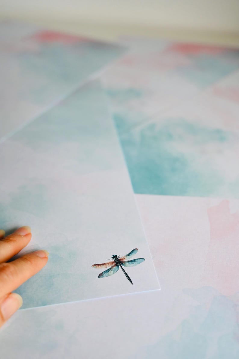Peut inclure: Une pile de papier &agrave; lettres blanc avec un motif aquarelle de nuages bleus et roses. Une libellule est imprim&eacute;e sur la feuille de papier sup&eacute;rieure.