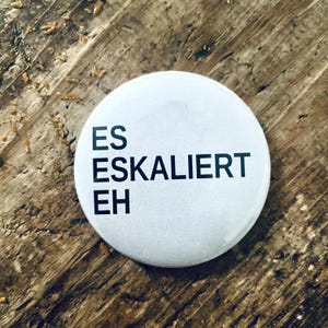 Könnte beinhalten: Ein weißer Button mit dem schwarzen Text "ES ESKALIERT EH". Der Button liegt auf einer strukturierten Holzoberfläche.