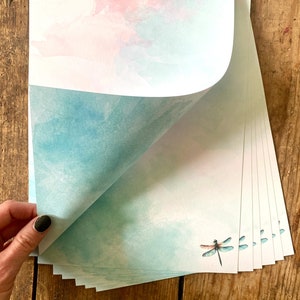 Peut inclure: Une pile de papier &agrave; lettres avec un fond aquarelle dans des tons de rose et de bleu. Le papier pr&eacute;sente une d&eacute;licate illustration de libellule.