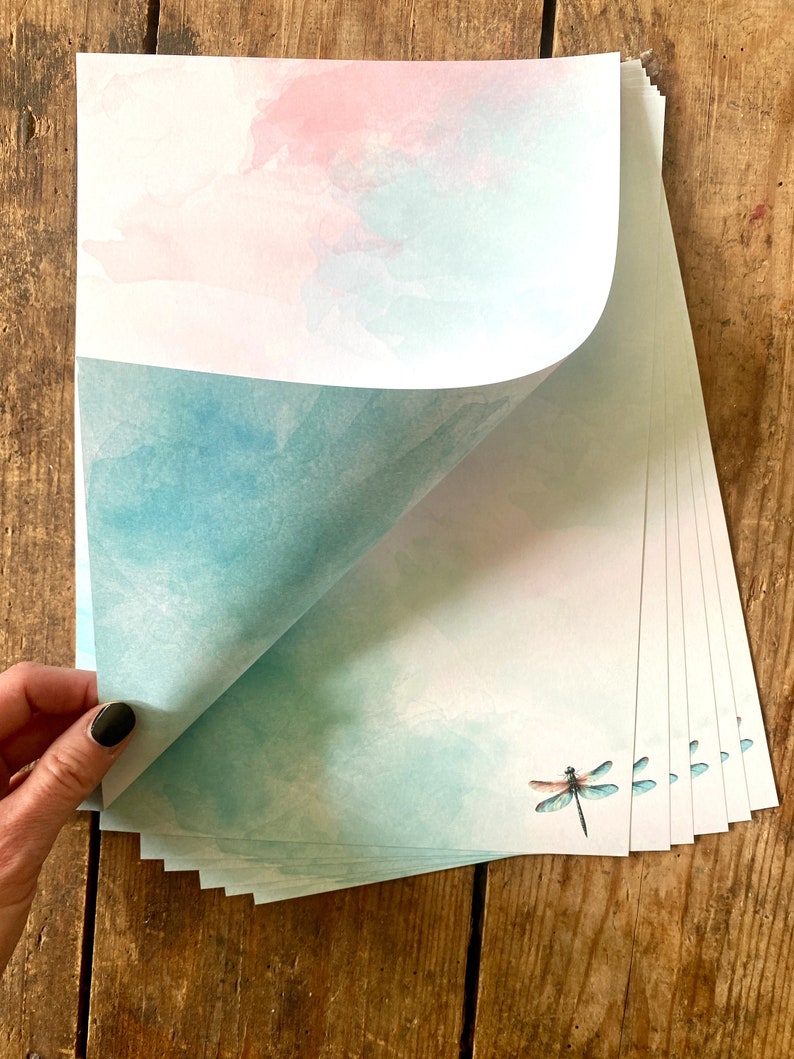 Peut inclure: Une pile de papier &agrave; lettres avec un fond aquarelle dans des tons de rose et de bleu. Le papier pr&eacute;sente une d&eacute;licate illustration de libellule.