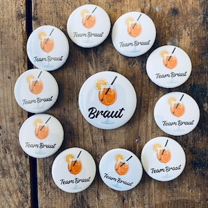 Pode incluir: Dez botões redondos brancos com uma bebida laranja e o texto "Team Braut" em preto. Um botão tem o texto "Braut" em preto.
