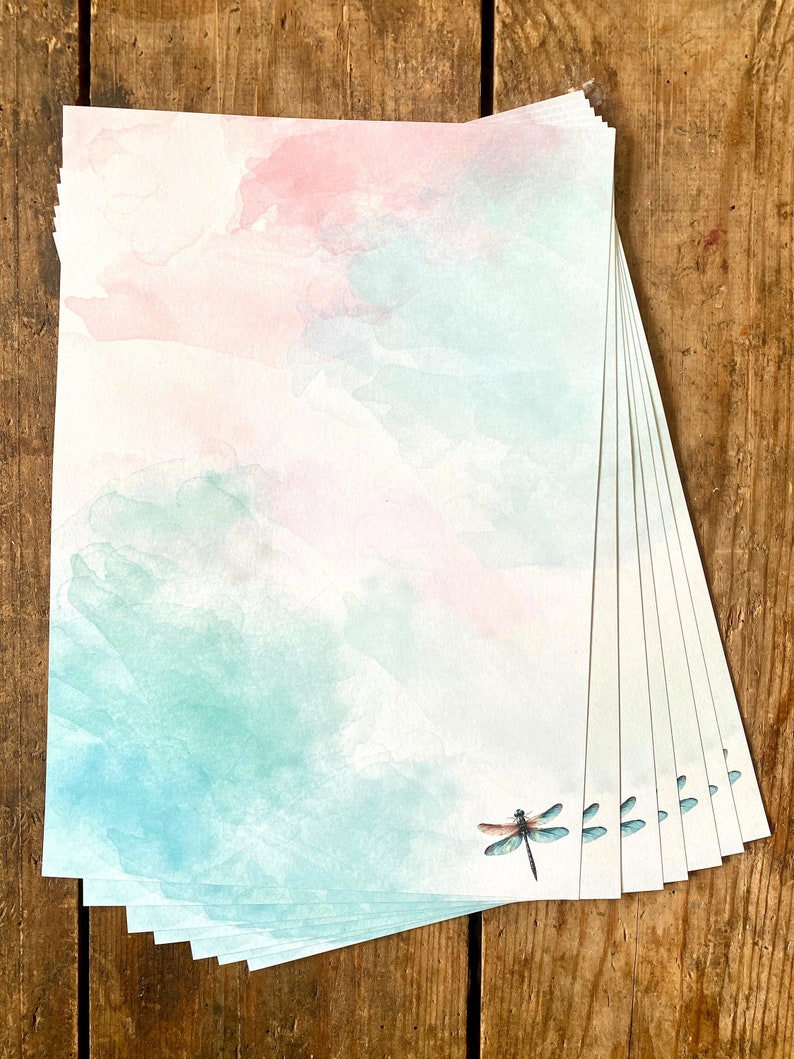 Peut inclure: Une pile de papier &agrave; lettres vierge avec un fond aquarelle dans des tons de rose et de bleu. Une libellule est imprim&eacute;e dans le coin inf&eacute;rieur droit du papier.