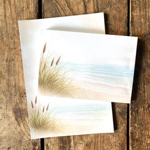Könnte beinhalten: Zwei leere Notizkarten mit einer Aquarell-Strandszene. Die Karten zeigen einen Sandstrand, das Meer und hohes Gras in Beige-, Blau- und Brauntönen. Die Karten liegen auf einer rustikalen Holzoberfläche.