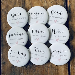 Peut inclure: Neuf boutons ronds blancs avec du texte noir et des cœurs roses. Le texte sur les boutons comprend des noms et des titres tels que "Gabi Bräutigammutter", "Siegfried Brautvater", "Gerd Bräutigamvater", "Sabine Brautmutter", "Julia Tranzengin Braut", "Nico Franzenge Bräutigam", "Lukas Bruder Bräutigam", "Tim Trauzenge Brantigam", et "Torsten Bruder Braut".