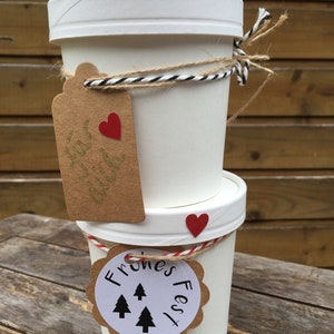 K&ouml;nnte beinhalten: Zwei wei&szlig;e Pappbecher mit braunen Papieretiketten, die mit Schnur zusammengebunden sind. Der obere Becher hat ein Etikett mit einem roten Herzen und dem Text "Festive Delight". Der untere Becher hat ein Etikett mit dem Text "Frones Fest" und drei schwarzen Tannenb&auml;umen.