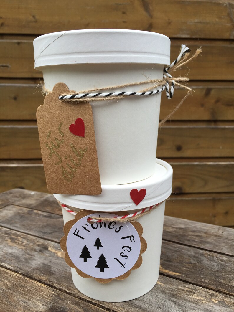 K&ouml;nnte beinhalten: Zwei wei&szlig;e Pappbecher mit braunen Papieretiketten, die mit Schnur zusammengebunden sind. Der obere Becher hat ein Etikett mit einem roten Herzen und dem Text "Festive Delight". Der untere Becher hat ein Etikett mit dem Text "Frones Fest" und drei schwarzen Tannenb&auml;umen.