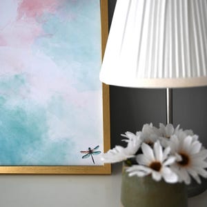Peut inclure: Une peinture &agrave; l'aquarelle encadr&eacute;e d'une libellule sur fond de ciel rose et bleu. La peinture est sur une surface blanche &agrave; c&ocirc;t&eacute; d'un abat-jour blanc et d'un vase de fleurs blanches.
