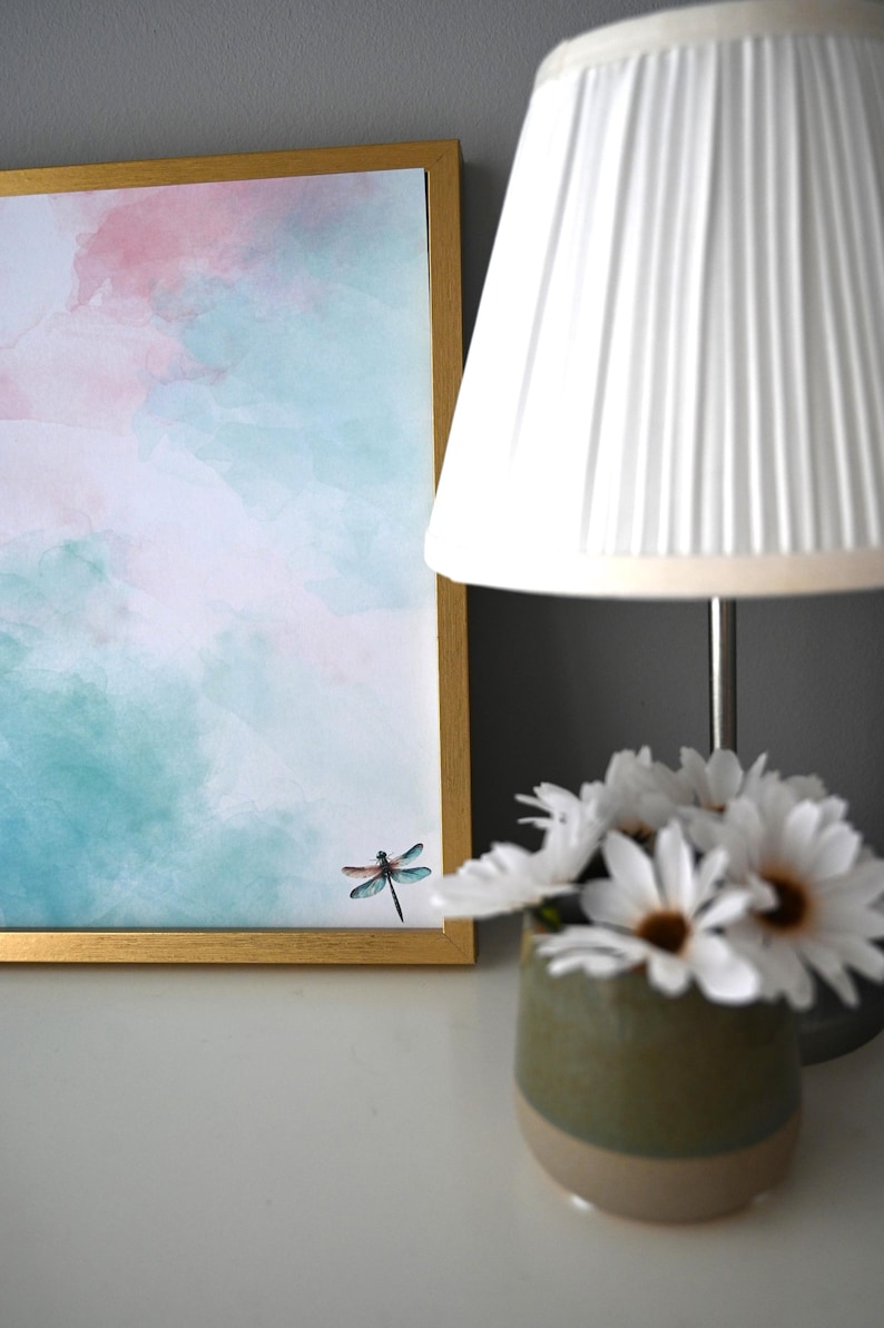 Peut inclure: Une peinture &agrave; l'aquarelle encadr&eacute;e d'une libellule sur fond de ciel rose et bleu. La peinture est sur une surface blanche &agrave; c&ocirc;t&eacute; d'un abat-jour blanc et d'un vase de fleurs blanches.
