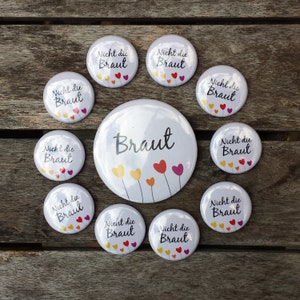 Könnte beinhalten: Neun weiße runde Knöpfe mit dem Text "Nicht die Braut" und einer Grafik mit Herzen in verschiedenen Farben. Ein größerer weißer runder Knopf mit dem Text "Braut" und einer Grafik mit Herzen in verschiedenen Farben.