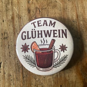 May include: A round button with the text "TEAM GLÜHWEIN" in brown. The button features a graphic of a mug of mulled wine with a cinnamon stick, orange slice, and star anise. The background is cream-colored.