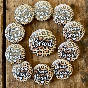 Può includere: Una collezione di bottoni con stampa leopardata con le parole "Team Braut" e "Braut" in corsivo. Ogni bottone ha un cuore bianco. I bottoni sono disposti su una superficie di legno.