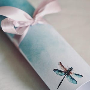 Peut inclure: Une impression enroul&eacute;e avec un fond aquarelle bleu clair et un ruban rose nou&eacute; autour. Une libellule est imprim&eacute;e sur le devant de l'impression.