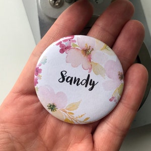 Könnte beinhalten: Ein weißer runder Knopf mit einem Blumendesign in Rosa-, Gelb- und Grüntönen. Der Name "Sandy" ist in schwarzer Kursivschrift geschrieben.