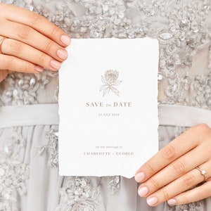 May include: A white, textured 'Save the Date' card with a floral illustration and the date '10 July 2024'. The card is held against a backdrop of a silver, embellished dress, suggesting a wedding theme.