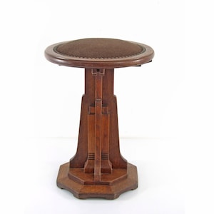 May include: A vintage wooden stool with a round, brown upholstered seat. The stool has a dark brown, geometric base and a central support with vertical and horizontal design elements. The seat is approximately 15 inches in diameter.