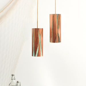 May include: A dining room table setting with a white tablecloth, black plates, a woven tray, and two copper pendant lights with a green and brown pattern. A wicker chair is in the background.