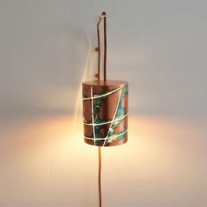 Op de afbeelding: Een cilindrische wandlamp met een metallic koperen afwerking, vervaagde blauwgroene vlekken en dunne, kruisende witte lijnen. De lamp is bevestigd aan een dunne koperkleurige stang en verspreidt een warme gloed aan de boven- en onderkant.