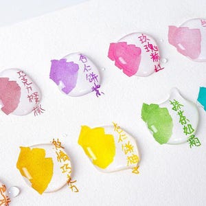 May include: A collection of circular white paint swatches on a white surface, each featuring a distinct color including orange, pink, purple, yellow, and green, with Chinese characters written in ink next to each individual paint sample.