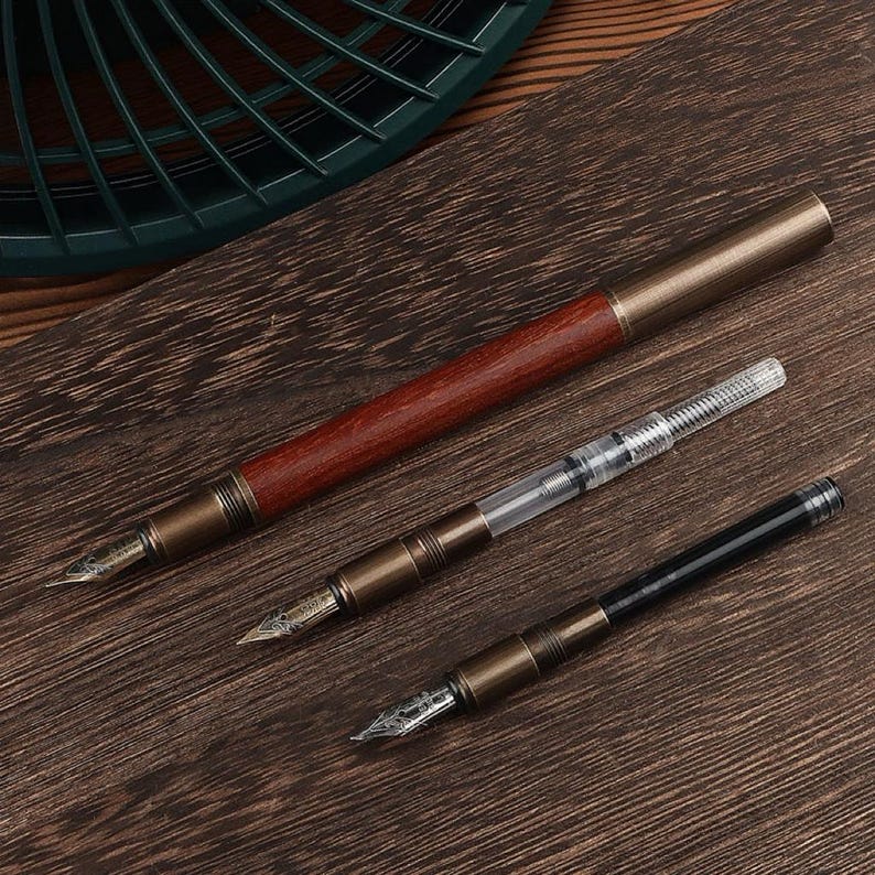 May include: Three fountain pens with different designs. One pen has a dark wood barrel and a brass-colored cap and accents. The other two pens have brass-colored accents and black or clear barrels. All pens have nibs.