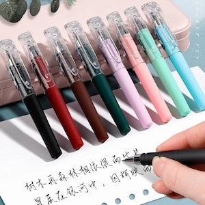 Peut inclure: Ensemble de stylos colorés avec des capuchons transparents et de l'encre noire. Les stylos sont disponibles en noir, rouge, marron, vert, violet, rose, vert clair et bleu. Une main écrit sur une feuille de papier.