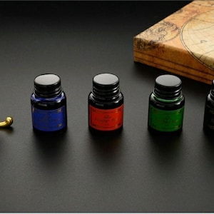 Op de afbeelding: Vier kleine glazen inktflesjes met zwarte schroefdoppen, elk voorzien van een gekleurd etiket in blauw, rood, groen en zwart. Naast de flesjes staat een bruine vulpen op een goudkleurige metalen standaard en een doos met een vintage kaart.