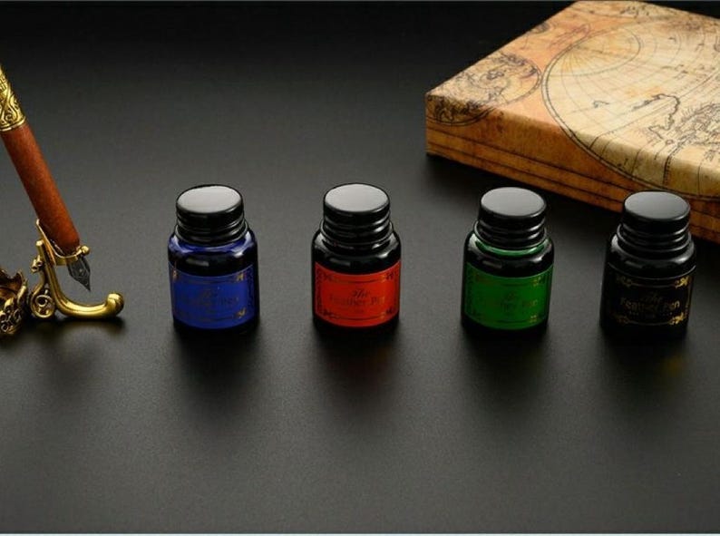 Op de afbeelding: Vier kleine glazen inktflesjes met zwarte schroefdoppen, elk voorzien van een gekleurd etiket in blauw, rood, groen en zwart. Naast de flesjes staat een bruine vulpen op een goudkleurige metalen standaard en een doos met een vintage kaart.