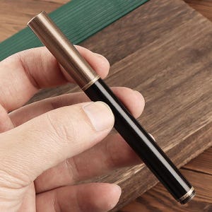 May include: A sleek, black and gold pen with a metallic finish. The pen has a dark barrel and a gold-colored cap and tip. The pen is held in a hand against a wooden background.