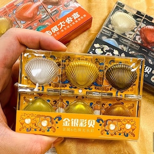 May include: A clear plastic box containing six seashell-shaped makeup products in various metallic colors: gold, bronze, and silver. The box is held in a hand and is labeled with Chinese characters. Other boxes with similar products are visible in the background.