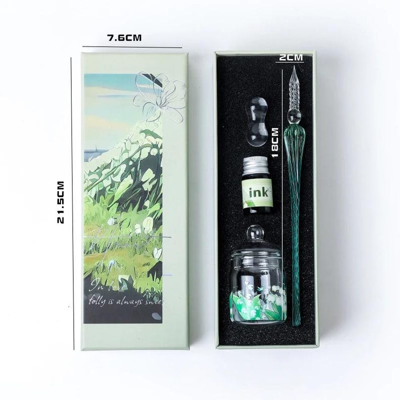 K&ouml;nnte beinhalten: Ein Kalligraphie-Set in einer Geschenkbox. Das Set enth&auml;lt einen gr&uuml;nen Glas-Dip-Pen (18cm), eine Tintenflasche, ein Tintenfass und einen kleinen Glas-Tintenhalter. Die Box ist 21,5cm hoch und 7,6cm breit, mit floralem Design und dem Text "Folly is always sweet."