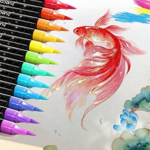 Op de afbeelding: Een set kleurrijke TrueColor brush pennen naast een gedetailleerde illustratie van een rode en roze koi vis. De pennen zijn gerangschikt in een regenboog van kleuren, waaronder rood, oranje, geel, groen, blauw en paars.