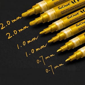 Op de afbeelding: Gouden acrylverfmarkers met verschillende puntmaten, waaronder 2,0 mm, 1,0 mm en 0,7 mm, worden getoond op een zwarte ondergrond. De markers, met het label "Touchmark", zijn ontworpen voor diverse oppervlakken.