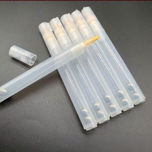 Peut inclure: Un ensemble de six stylos cosmétiques en plastique translucide avec des embouts pinceaux. Chaque stylo présente un corps cylindrique transparent contenant une petite bille de mélange en métal. Un stylo est montré sans son capuchon.