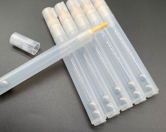 May include: A set of six translucent plastic cosmetic pens with brush tips. Each pen features a clear cylindrical body containing a small metal mixing ball inside. One pen is shown with its matching clear cap removed, revealing a beige brush tip.