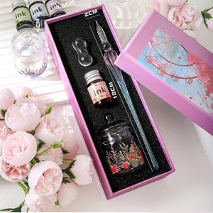 K&ouml;nnte beinhalten: Ein Kalligraphie-Set in einer rosa Box. Das Set enth&auml;lt einen Glasstift (18 cm), eine Tintenflasche, ein Tintenfass und eine Stifthalterung. Die Tintenflasche ist mit "ink" beschriftet. Die Box ist ge&ouml;ffnet und zeigt den Inhalt vor schwarzem Hintergrund. Die Box ist 21,5 cm lang.