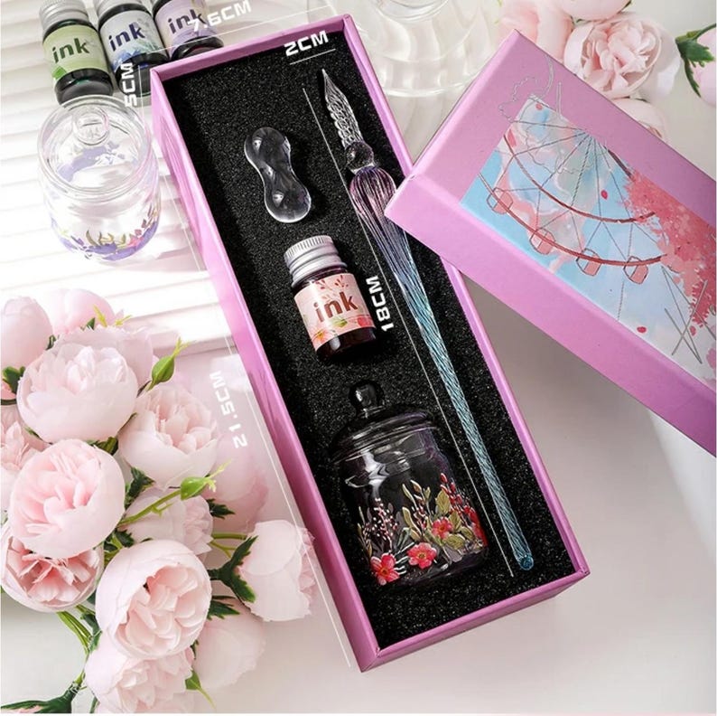 K&ouml;nnte beinhalten: Ein Kalligraphie-Set in einer rosa Box. Das Set enth&auml;lt einen Glasstift (18 cm), eine Tintenflasche, ein Tintenfass und eine Stifthalterung. Die Tintenflasche ist mit "ink" beschriftet. Die Box ist ge&ouml;ffnet und zeigt den Inhalt vor schwarzem Hintergrund. Die Box ist 21,5 cm lang.
